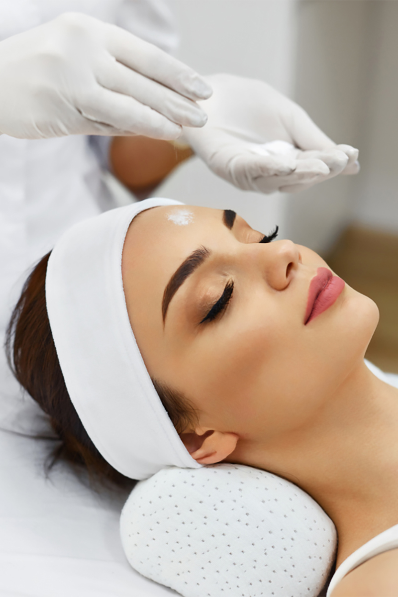 Serene woman receiving a holistic skin care treatment, with an aesthetician applying cream to her forehead.